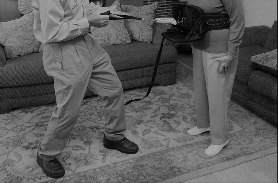 SST Anterior direction waist pull testing (rear stepping) set up with one end of the spring scale attached to the participant’s waist and the other end held by the examiner. A tether strap is secured via a belt at waist-level to both the participant and the examiner for participant safe. The figure also depicts a clear view of the participant’s feet and the presence of compliant surface to ensure participant safety.